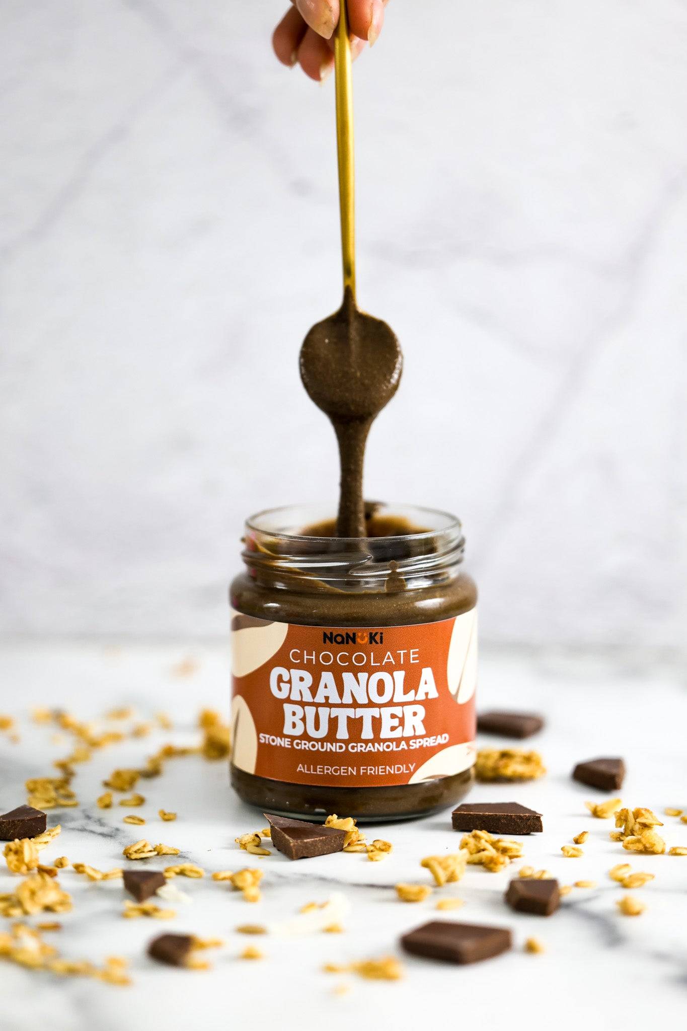 Granola Butter - Chocolate spread in a glass jar with a spoonful being lifted, surrounded by chocolate chunks and oats. Allergen-friendly dip in Food Items category.