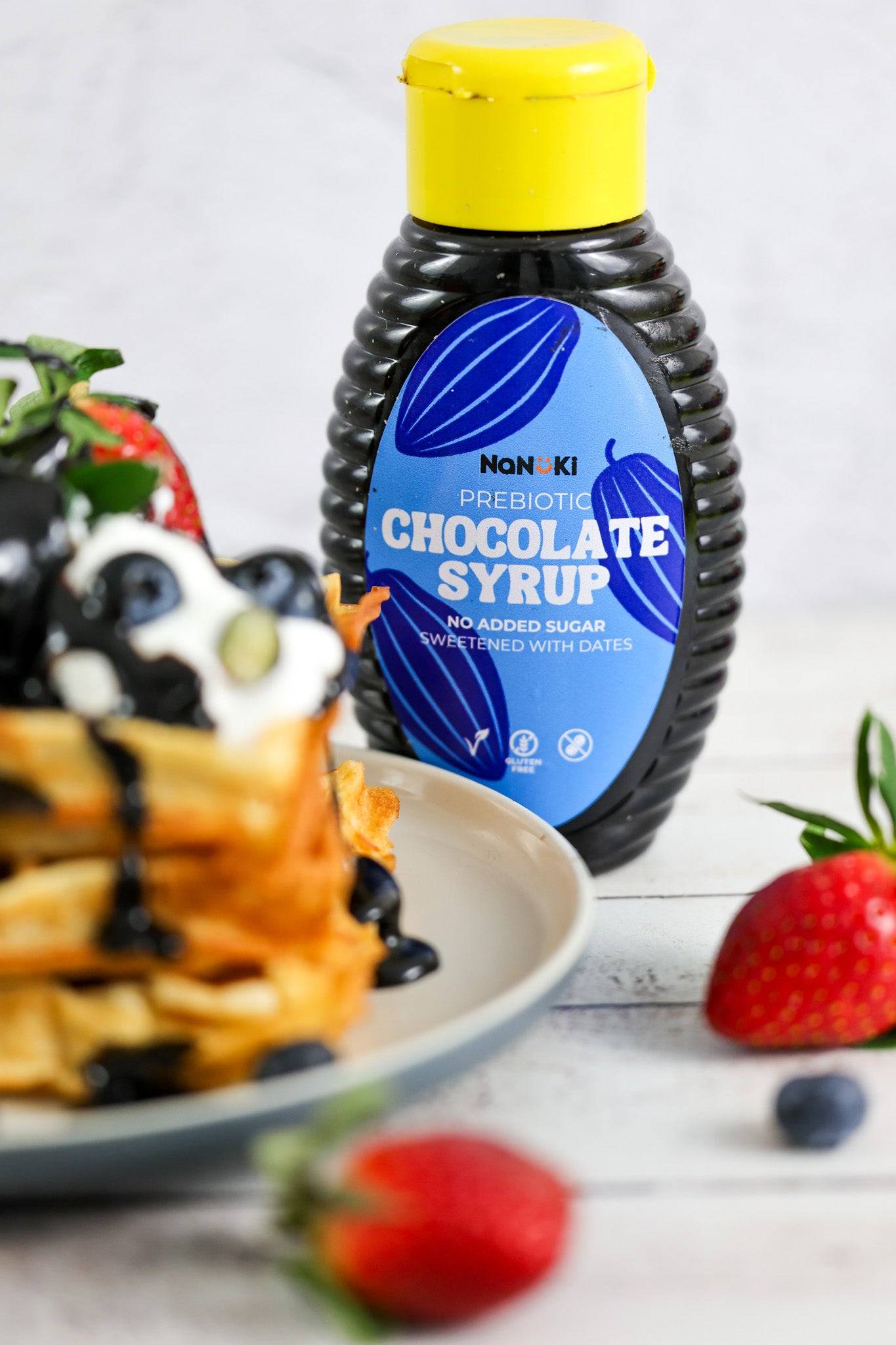 Nanoki Prebiotic Chocolate Syrup bottle with no added sugar, sweetened with dates, next to waffles topped with syrup, berries, and cream.