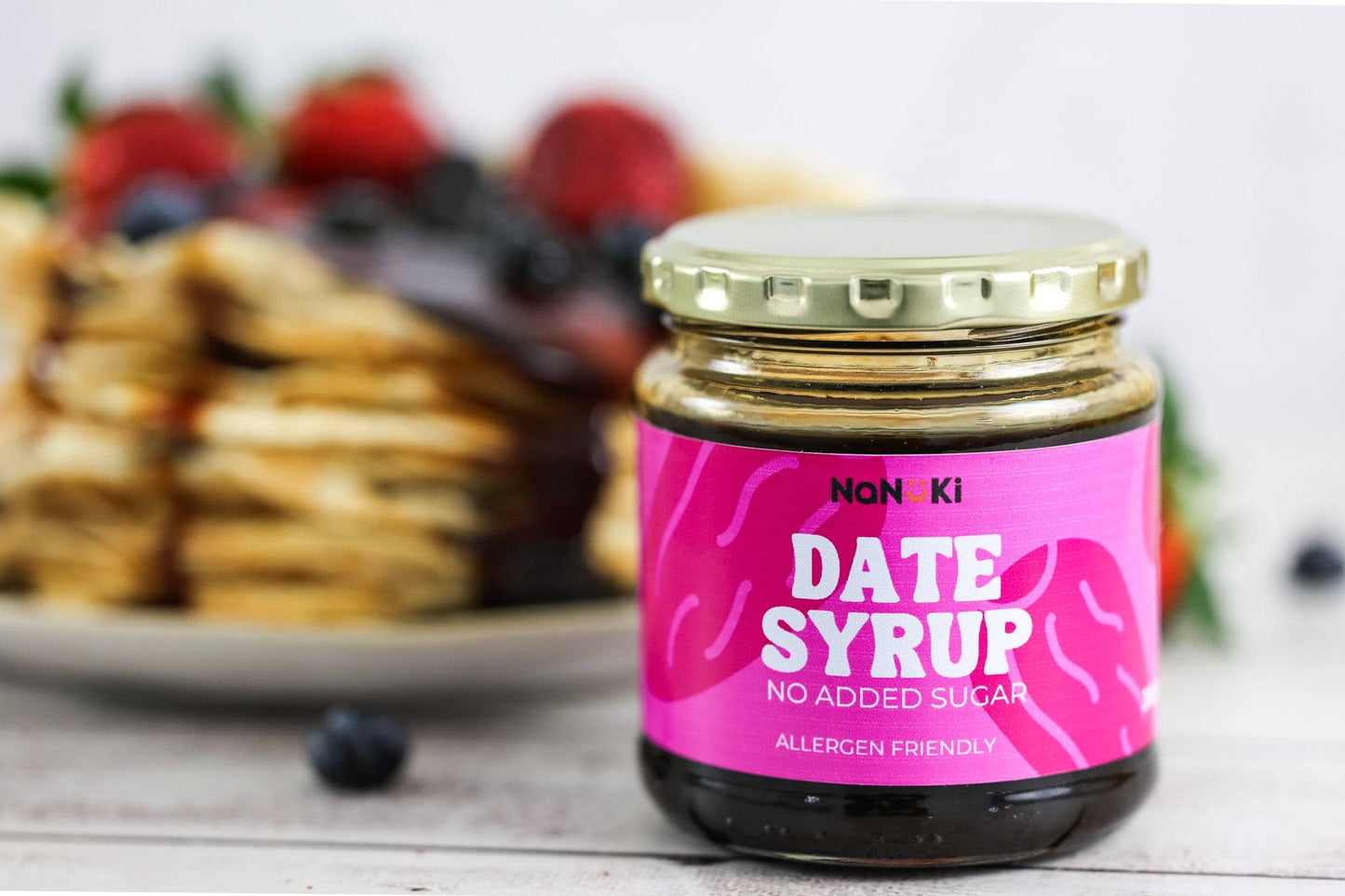 Date Syrup by NaNuKi in a glass jar with a gold lid, labeled allergen friendly and no added sugar, placed in front of a stack of pancakes with berries.