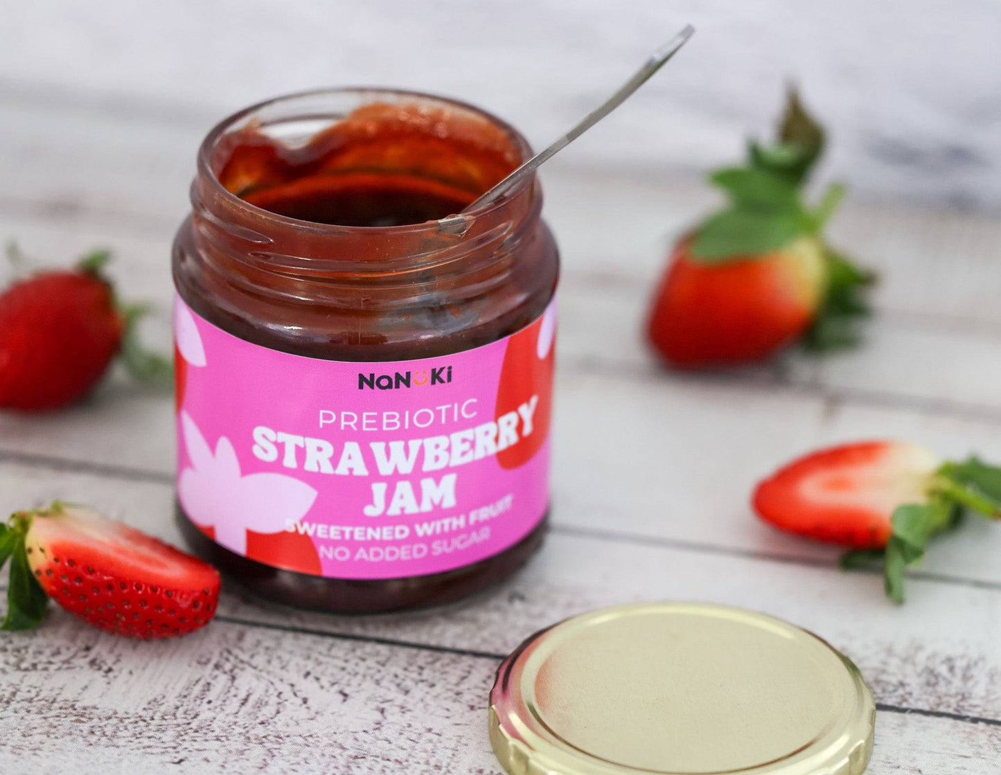 Prebiotic Strawberry Jam in an open glass jar with spoon, surrounded by fresh strawberries on a white wooden surface. No added sugar, fruit-sweetened spread.